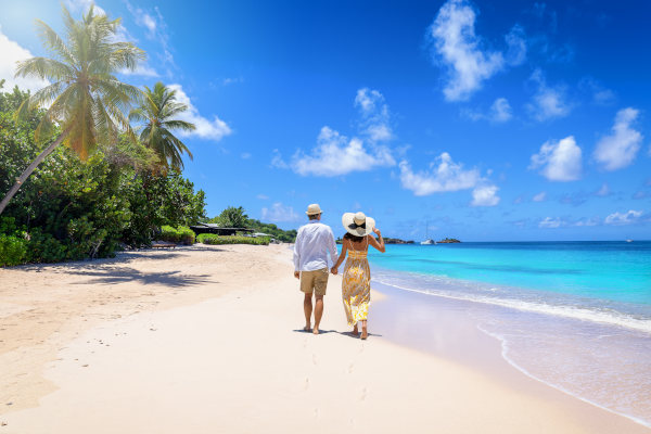 Pareja de turistas con sombrero y ropa de verano caminando por una playa del Caribe en un día soleado.