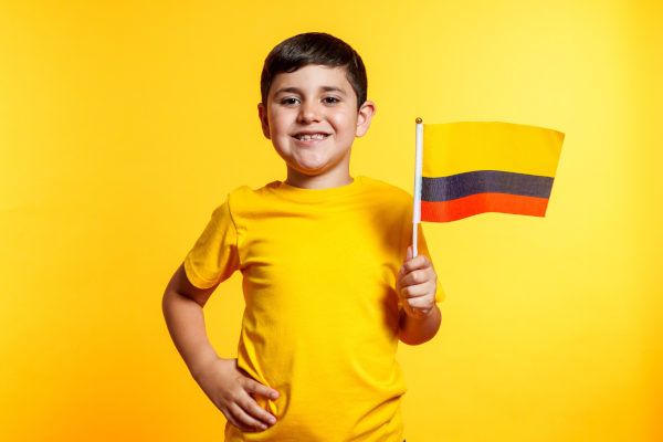 Niño sonriente sosteniendo la bandera de Colombia sobre un fondo liso amarillo.