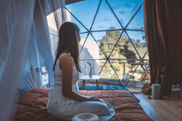 Mujer sentada en la cama de un domo de un glamping de lujo mirando hacia el paisaje verde exterior