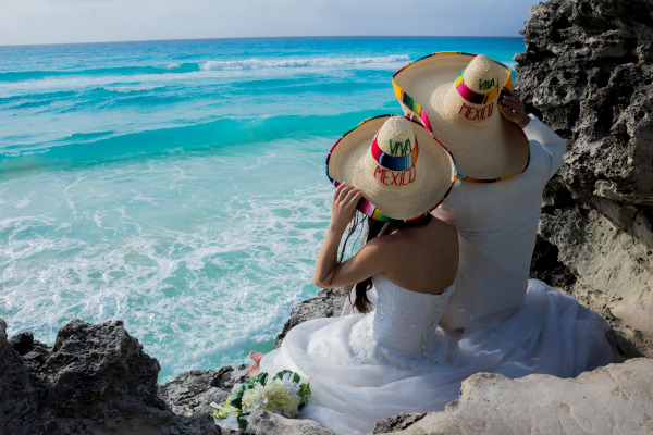 Pareja de recién casados vestidos de blanco sentados en un acantilado con vistas al mar turquesa y sombreros tradicionales mexicanos.