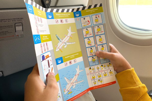 Vista de una tarjeta de seguridad con gráficos y texto en dos idiomas sostenida por un pasajero de avión.
