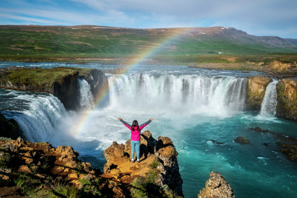 Turista con los brazos abiertos en un acantilado frente a la cascada Godafoss en un día soleado con arcoiris, Islandia.