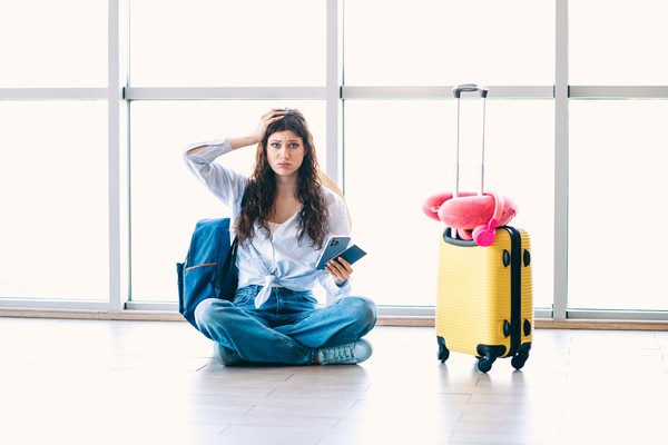 Mujer preocupada sentada en el suelo de una terminal de aeropuerto con maleta de viaje al lado.