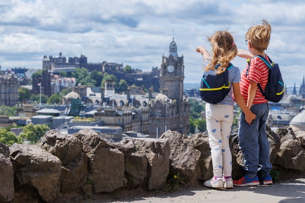 Dos niños con mochilas, tomados de la mano, apuntando a las vistas de la ciudad de Edimburgo, Escocia, desde un mirador rocoso.