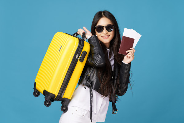 Mujer con lentes de sol cargando pasaporte, billete de avión y maleta amarilla con una sonrisa en un fondo azul claro liso.