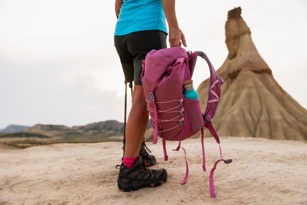Vista detallada de las piernas de una excursionista con botas de senderismo y mochila rosa en el desierto de Bardenas Reales con la icónica Castildetierra detrás, Navarra, España.