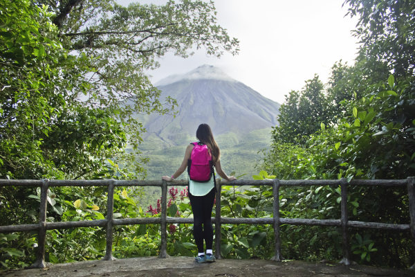Mujer turista con mochila fucsia en un mirador de espaldas a la cámara y de frente al Volcán Arenal en Costa Rica en un día nublado.