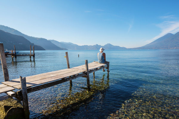 Hombre sentado en el borde de un muelle en el Lago Atitlán con vistas a las aguas transparentes y montañas de fondo, Guatemala.