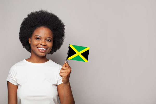 Mujer sonriente con cabello rizado sosteniendo la bandera de Jamaica sobre un fondo claro liso.