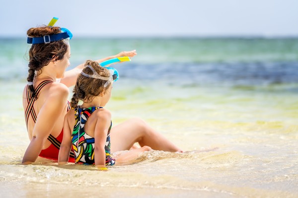 Madre e hija con máscaras de snorkel sentadas en la orilla del mar, mirando el horizonte.