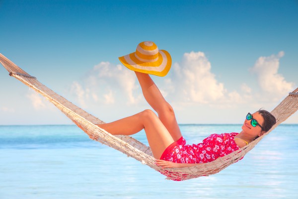 Mujer con lentes de sol y vestido playero tumbada en hamaca con el sombrero en el pie con aguas cristalinas de fondo.