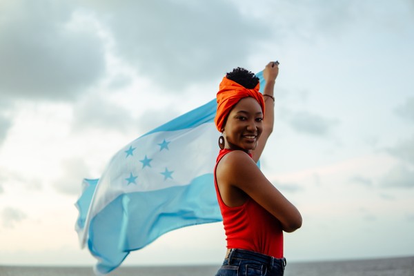 Mujer garífuna ondeando la bandera hondureña al viento en una playa en un día nublado.