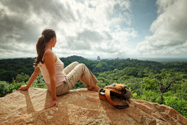 Mujer joven con mochila sentada en una roca en la altura mirando la vegetación y las ruinas de una antigua ciudad maya de Tikal, Parque Nacional Tikal, Guatemala.