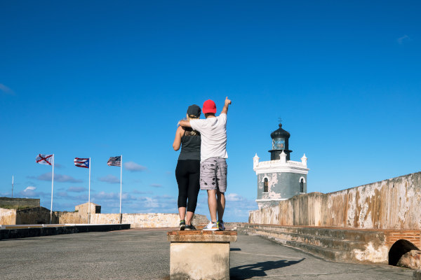 Pareja de turistas en una de las fortalezas de la Bahía de San Juan con las banderas de Puerto Rico y de Estados Unidos de fondo en un día soleado.