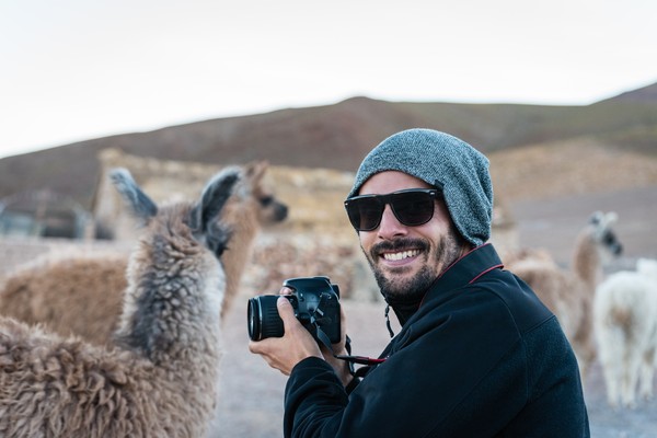Hombre turista con cámara fotográfica sonriendo mientras toma fotos de alpacas en el altiplano andino en Bolivia.