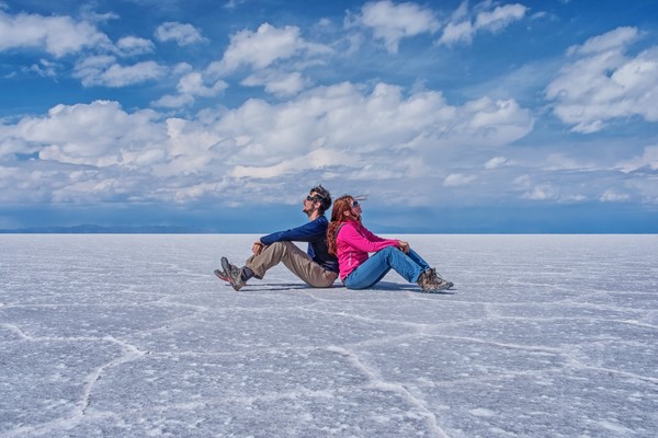 Pareja de viajeros sentados espalda contra espalda en el suelo del Salar de Uyuni, Bolivia, con nubes de fondo.