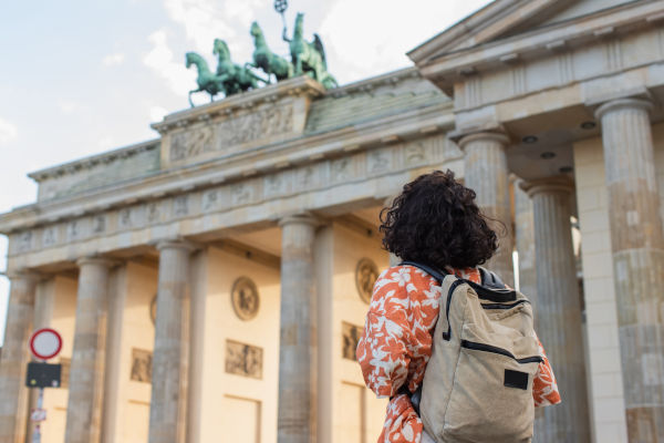 Vista trasera de una turista de cabello rizado con mochila, de pie frente a la Puerta de Brandeburgo en Berlín.