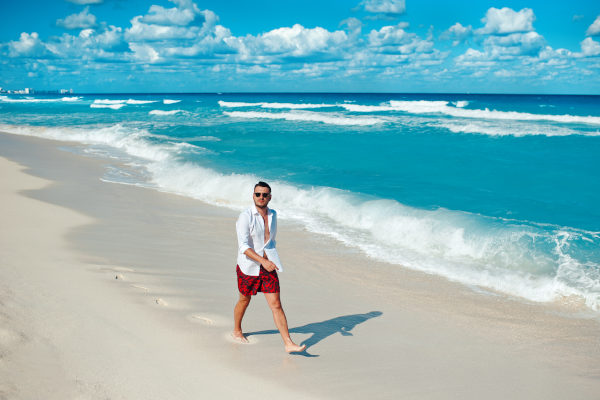 Joven turista masculino con camisa blanca y gafas de sol caminando solo en la playa de Cancún, México.