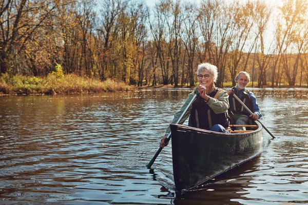 Pareja de ancianos sonrientes en un bote tipo kayak remando en un lago tranquilo con arboleda de fondo.