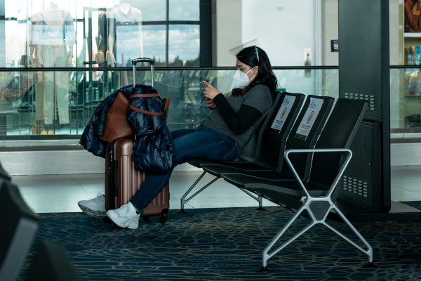 Mujer latina con mascarilla sentada en la terminal, usando su celular mientras espera junto a su equipaje.