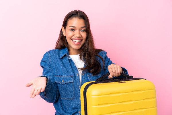 Mujer colombiana con maleta amarilla en mano, sonriente, sobre fondo rosa.