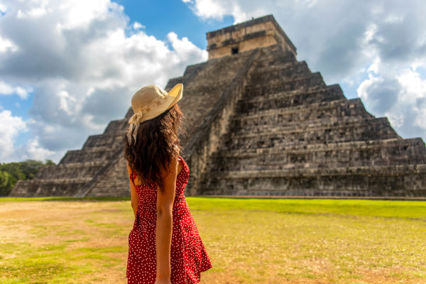 Mujer de espaldas con vestido rojo y sombrero contemplando una pirámide antigua en México bajo el sol.