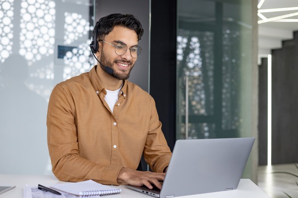 Hombre representante de atención al cliente trabajando en la laptop con una sonrisa y auriculares con micrófono puestos