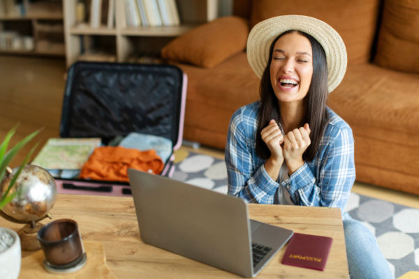 Mujer feliz planeando su viaje desde casa, con maleta abierta, pasaporte y laptop sobre la mesa.