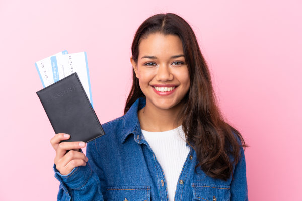 Mujer sonriente sobre fondo liso rosado mostrando pasaporte y boletos de avión