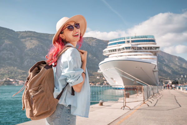 Mujer turista con sombrero y mochila sonriendo con entusiasmo cerca de un crucero detenido en un puerto en un día soleado