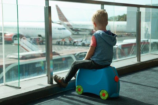 Niño sentado en una maleta de paseo, mirando por la ventana del aeropuerto a los aviones en la pista.