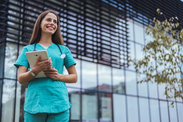 Médica con tablet y estetoscopio sonriendo en las afueras de un hospital.