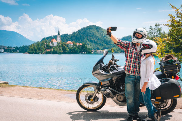Pareja de motociclistas se detiene a un costado de la carretera para tomarse una fotografía junto a un lago en un día soleado.