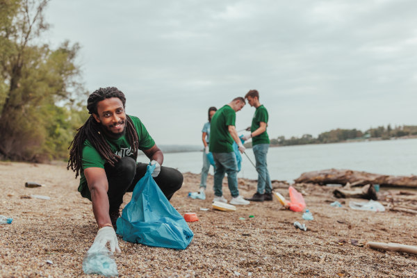 Voluntario ambientalista recoge basura en una playa contaminada mientras mira a cámara con una sonrisa.