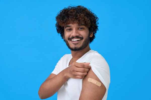 Hombre sonriente con barba y cabello enrulado arremangándose para mostrar en el brazo un apósito cubriendo la zona de una vacuna reciente.