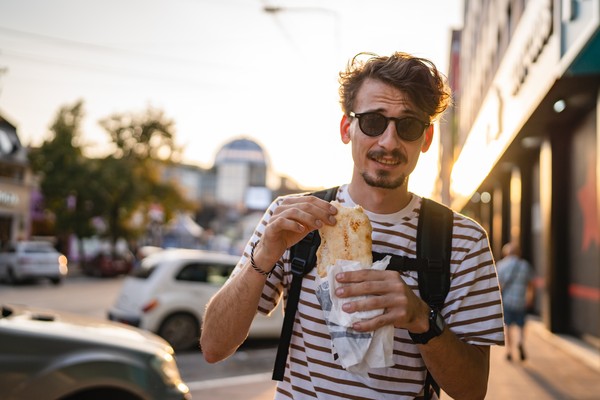 Hombre con mochila posando para la foto con comida callejera en la calle de una ciudad.