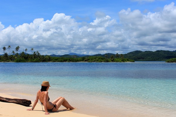 Mujer joven en traje de baño sentada en la arena remojando los pies en el mar en la playa de la isla  Pelícano, territorio indígena Kuna, San Blas, Panamá.