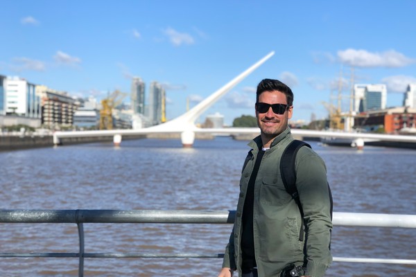 Hombre turista feliz posando para una foto en una barandilla con el Río de la Plata y el Puente de la Mujer de fondo en un día soleado en Puerto Madero, Buenos Aires, Argentina.
