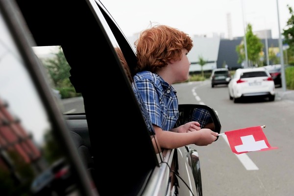 Niño asomado por la ventanilla delantera de un auto con una bandera pequeña de Suiza.