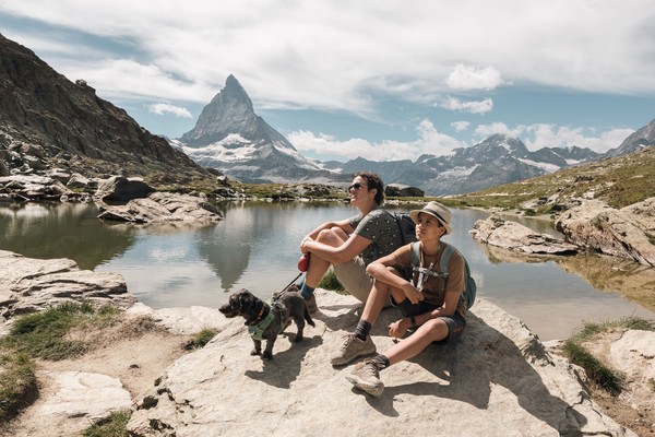 Dos hombres sentados en una roca con un perro disfrutando de la belleza de las montañas junto a un lago con el pico Matterhorn de fondo en Suiza.