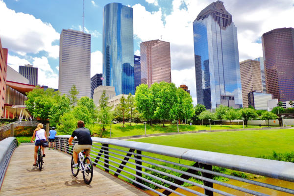 Grupo de ciclistas atravesando un puente de madera en el Parque Buffalo Bayou, con una hermosa vista del centro de Houston en un día de verano.