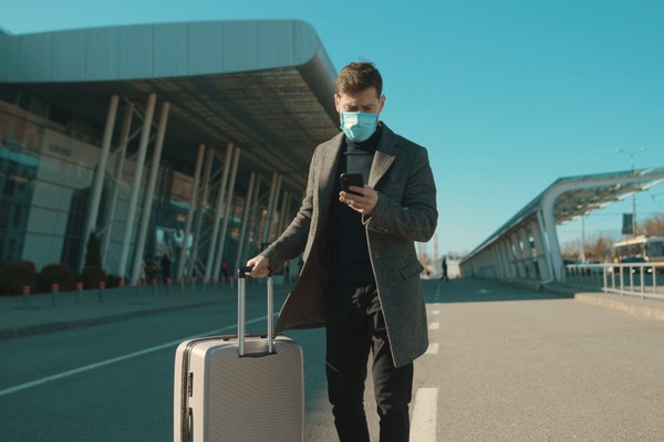 Hombre con mascarilla y maleta de pie mirando el teléfono móvil en la entrada de un aeropuerto con preocupación.