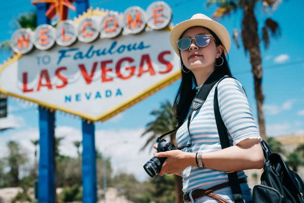 Mujer turista con lentes de sol y sombrero sosteniendo cámara fotográfica con el famoso letrero de Welcome to Fabulous Las Vegas de fondo.