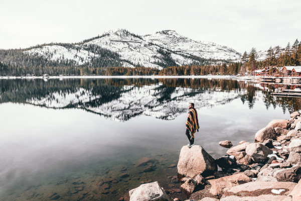 Hombre con poncho parado en una roca en la orilla del Lake Tahoe en invierno con montañas nevadas de fondo.