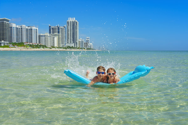 Dos niños con lentes de sol nadando con un flotador en las aguas cristalinas de Miami con la playa y los edificios de la ciudad de fondo