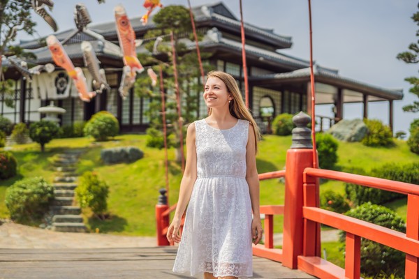 Mujer viajera sonriente con vestido blanco caminando por un puente de madera con edificio tradicional japonés de fondo.