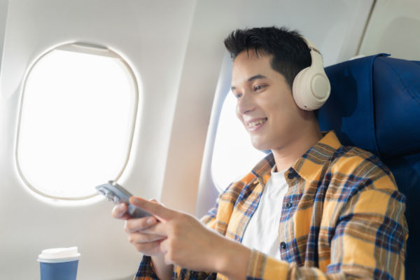 Joven viajero en un avión, sonriendo mientras usa su celular y escucha música con audífonos,