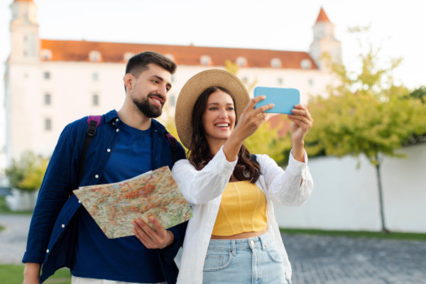 Pareja joven viajera tomándose una selfie con el celular mientras consultan un mapa.