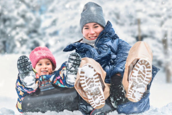 Madre e hija jugando en la nieve, deslizándose en un trineo, con rostros sonrientes y ropa de invierno.