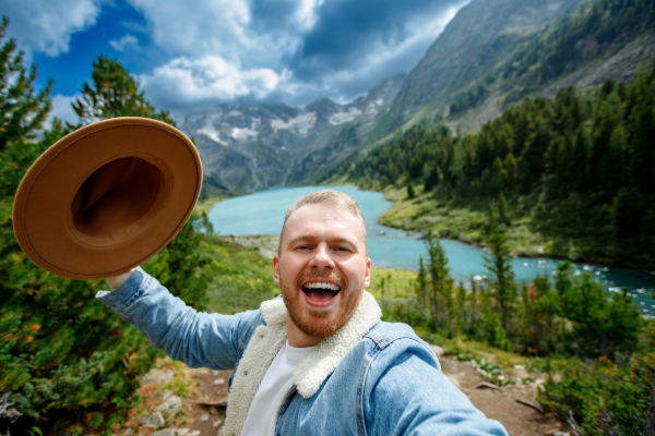 Joven sonriente levantando su sombrero frente a un lago rodeado de montañas y bosques.